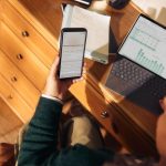 Person holding phone near laptop and tablet on desk